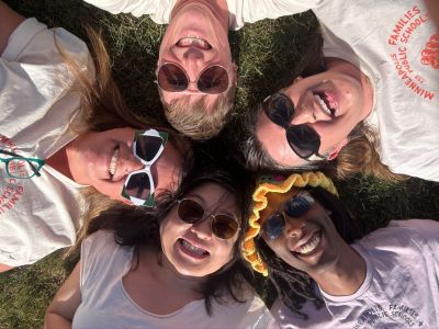 A group of five people in sunglasses laying on the ground with their heads together in a circle smiling up at the viewer above.