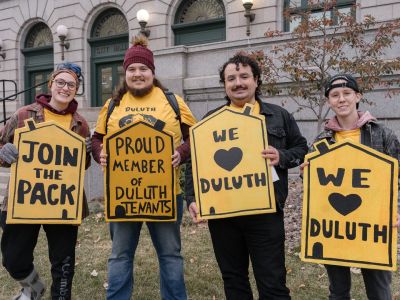 A group of four smiling members of Duluth Tenants standing in front of a building, holding demonstration signs in bright yellow supporting their community cause.