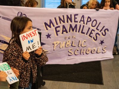 A young child holding a sign that says "Invest in me!", in front of a large hand-painted banner that reads "Minneapolis Families for Public Schools".
