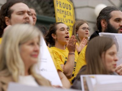 A closely cropped photograph of a large group of Duluth community members standing together outside a building holding signs and joyfully raising their voices together. Yellow is a prominent color in both clothing and signs they are holding.