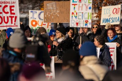 A large group of Minnesota community members gathered together in demonstration holding colorful, bold messaged signs supporting community and ICE Out of Minnesota. The crowd dressed in winter coats and hats is addressed by a speaker standing at the center holding a microphone.