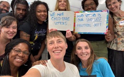 A group of parents posing for a picture, some holding signs that say "We are child care voters..." and "And voting is our smallest political act!"