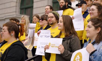 A closely cropped photograph of a large group of Duluth community members standing together outside a building holding signs. Yellow is a prominent color in both clothing and signs they are holding.