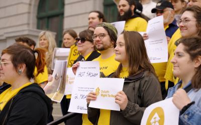 A closely cropped photograph of a large group of Duluth community members standing together outside a building holding signs. Yellow is a prominent color in both clothing and signs they are holding.