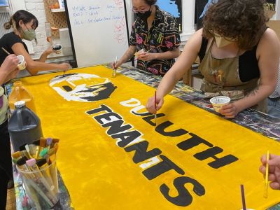 A group of Duluth Tenant team members working together at a table painting a yellow and black sign with the text Duluth Tenants and their log being filled in.