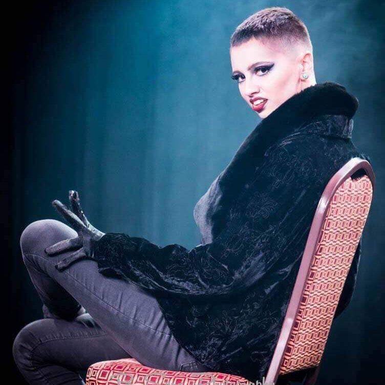 Burlesque performer Justin Uranus poses for the camera. They are seated on a red chair and wearing a black fur coat and stage makeup, with a blue curtainin the background. 