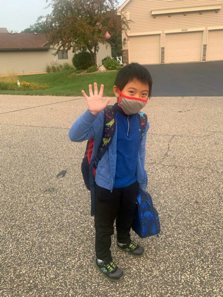 A photo of author LyLy's brother Leland, age 7,standing on a street in front of a house. Leland is wearing a blue jacket and a face mask, and waving at the camera.