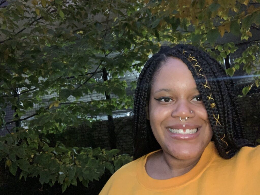 Cierra Brown smiles at the camera in an outdoor setting. She's wearing a yellow shirt. Sunshine filters through trees in the background.