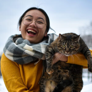 LyLy Vang-Yang holds her cat Bella up and laughs at the camera. Behind them is the blue sky and