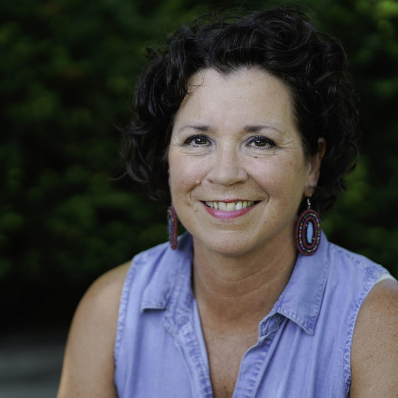 A photograph of State Representative and Senator-Elect Mary Kunesh-Podein smiling. She wears a light blue top. Dark greenery can be seen in the background of the outdoor setting.