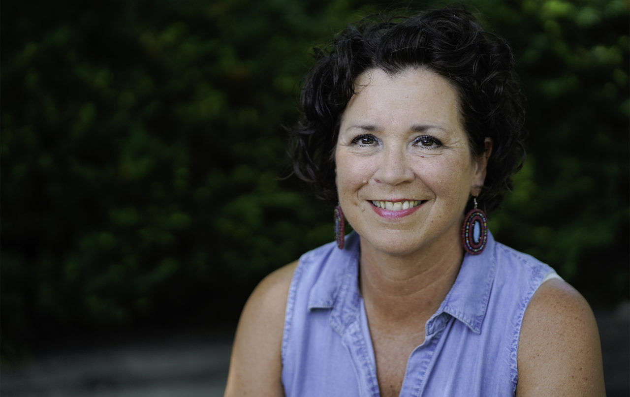 A photograph of State Representative and Senator-Elect Mary Kunesh-Podein smiling. She wears a light blue top. Greenery can be seen in the background of the outdoor setting.