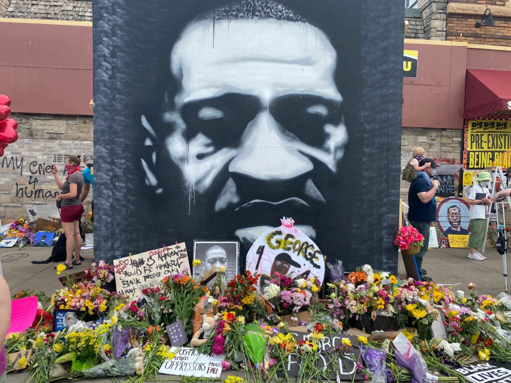 Photo: Black and white mural of George Floyd's face at 38th & Chicago (George Floyd Square). Gifts and flowers from visitors to the memorial cover the ground surrounding the mural. 