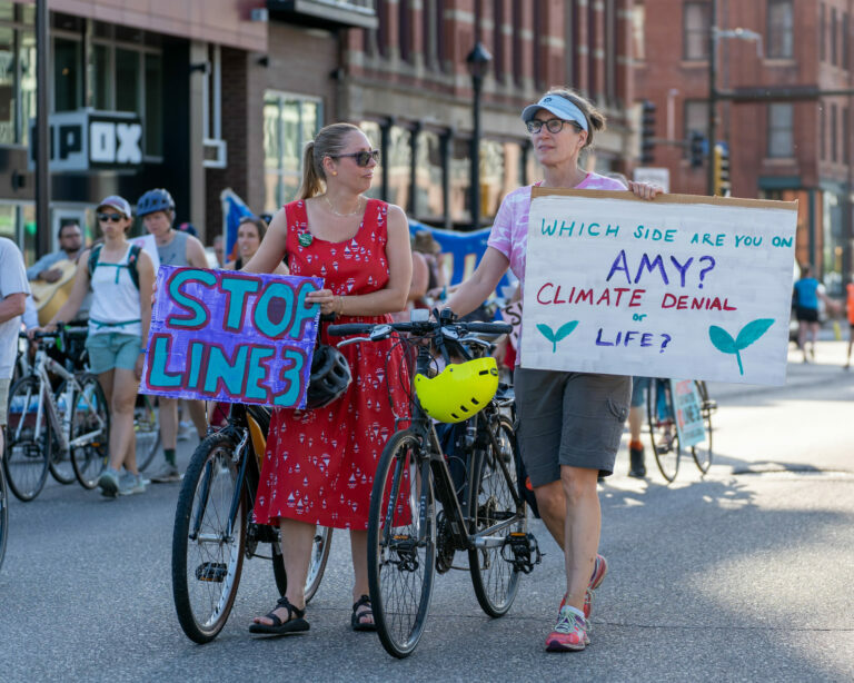 Protesters hold signs. One says "Which side are you on, Amy?"