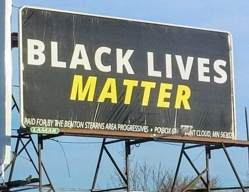 A billboard with white and yellow text on a black background: "Black Lives Matter. Paid for by the Benton Stearns Area Progressives - PO Box (not visible) Saint Cloud, MN 56302