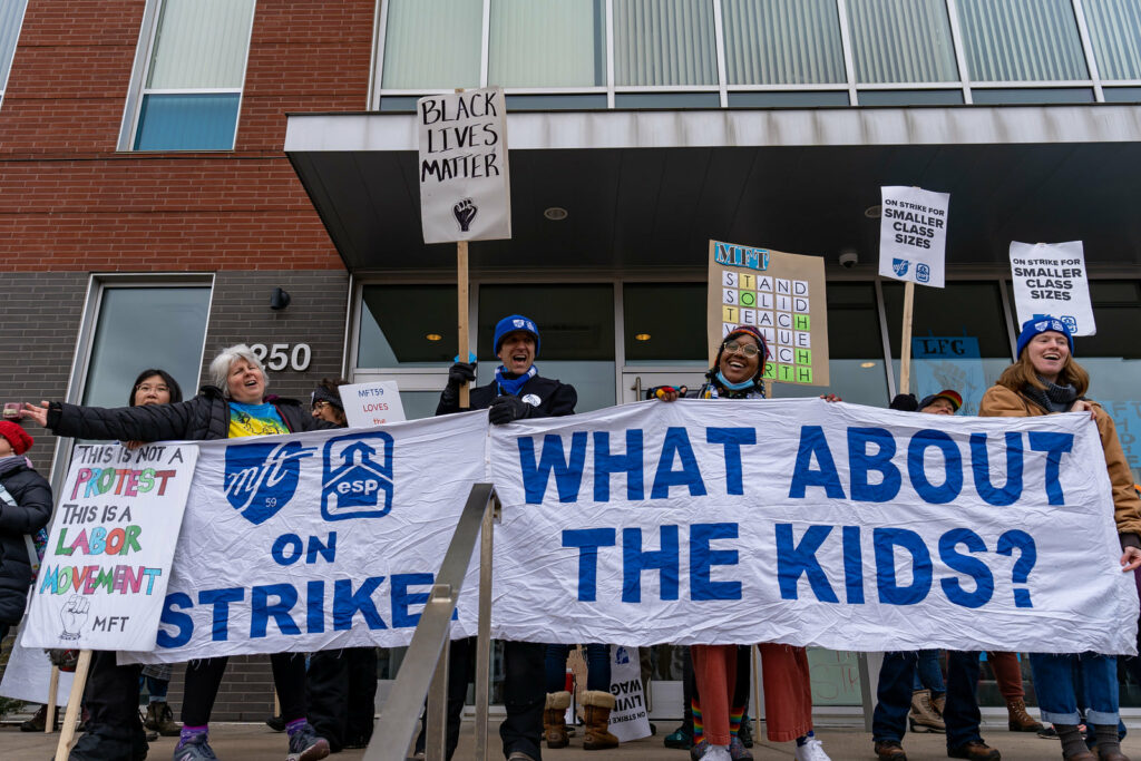 A multiracial group of educators stand in front of an education building holding picket signs and a banner that reads "MFT, ESP on STRIKE... WHAT ABOUT THE KIDS?"