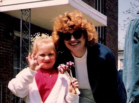 Photo of a red-haired woman and young blonde girl wearing a flower crown and holding a small bouquet of flowers, standing in front of an apartment building in the spring.