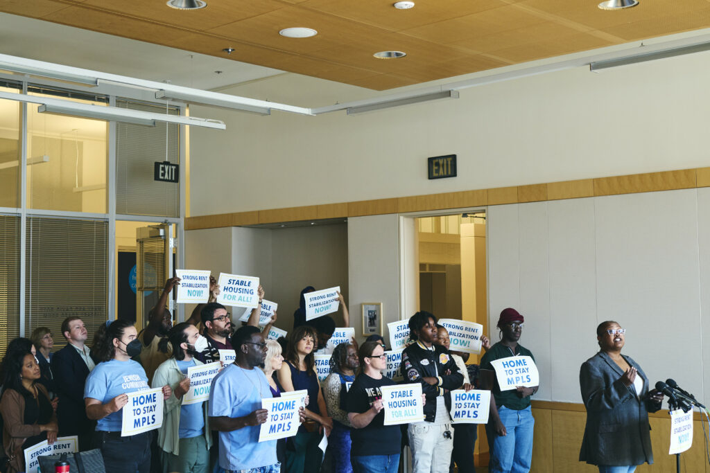 Photo from Home to Stay's Sept. 14, 2022 press conference: Yolanda Roth stands at a podium. Behind her, a diverse group of community members hold signs that say "Home to Stay MPLS" and "Stable Housing For All."