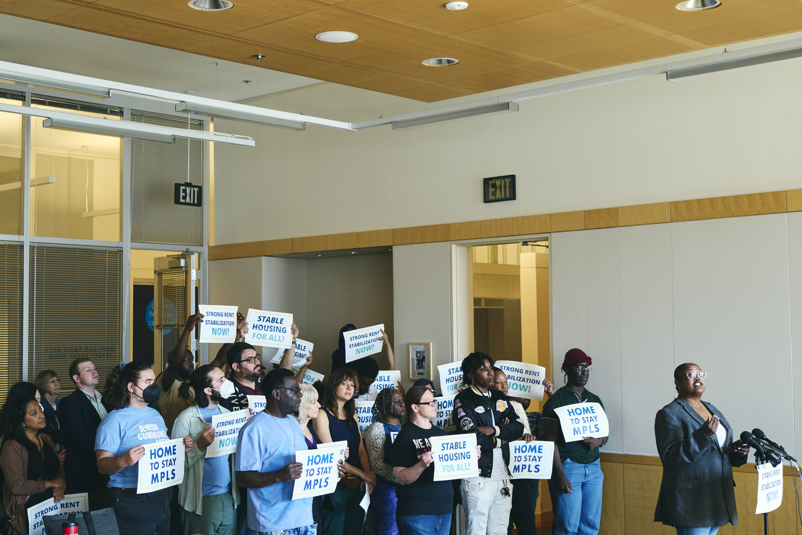 Photo from Home to Stay's Sept. 14, 2022 press conference: Yolanda Roth stands at a podium. Behind her, a diverse group of community members hold signs that say "Home to Stay MPLS" and "Stable Housing For All."