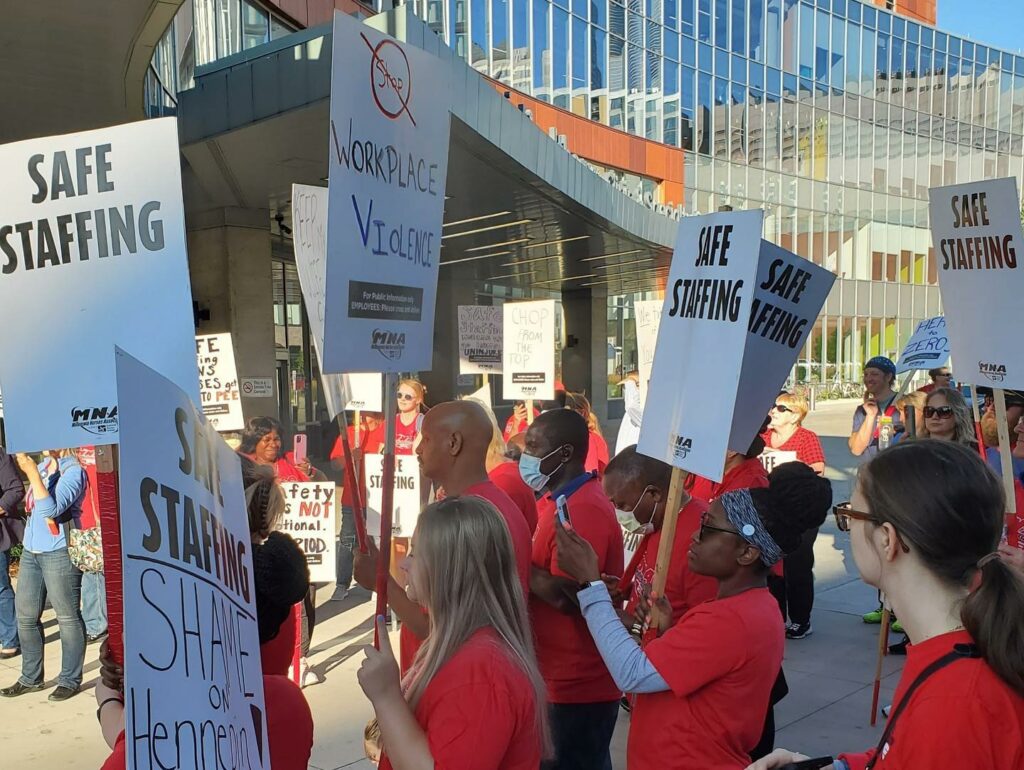 A group of nurses in red shirts hold signs that say "safe staffing" at a Minnesota Nurses Association press conference in August, 2022