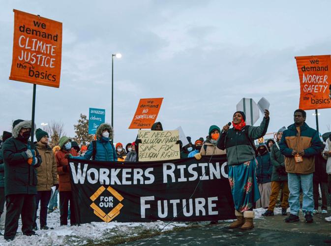 Amazon workers and community members rally, holding signs and a banner that reads "Workers Rising for our Future." Khali Jama speaks into a microphone.